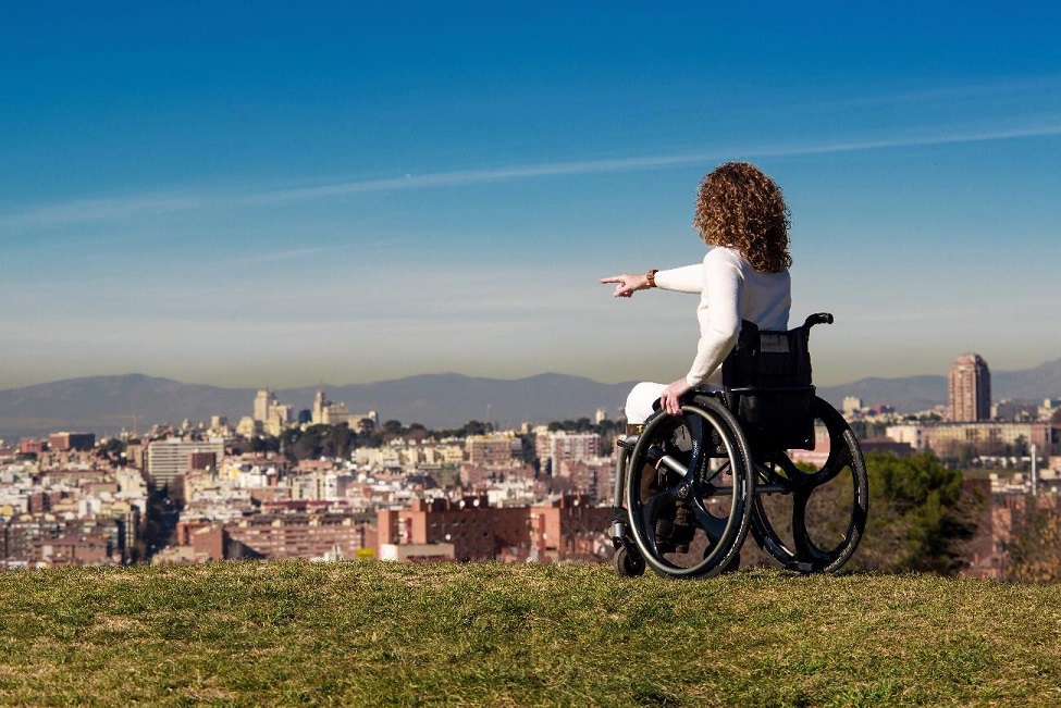 Imagen de una chica en silla de ruedas, señalando una ciudad, desde lo alto de una cima